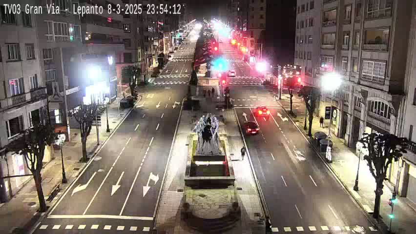 A nighttime, city street scene shows a multi-lane road with sparse traffic, buildings lining either side, and a monument in the center of the intersection.