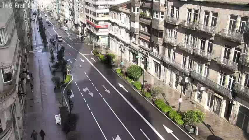 A wet city street lined with multi-story buildings, sparsely populated with pedestrians and a few cars.