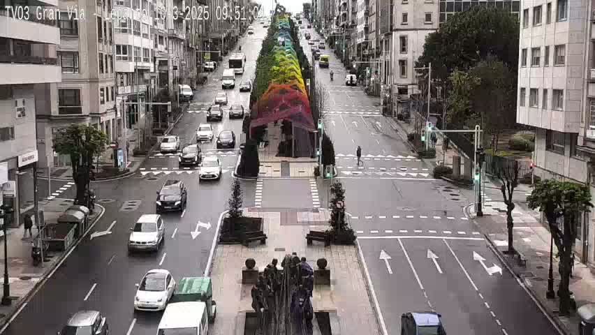 A rainy city street scene shows several cars at an intersection with a pedestrian walkway decorated with a rainbow-colored canopy.