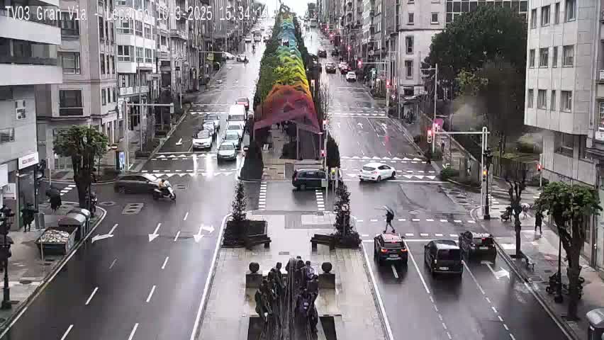 A rainy city street scene shows several cars and pedestrians navigating a multi-lane road with a colorful median.