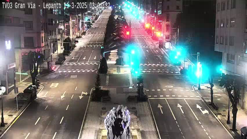 A mostly empty, multi-lane city street at night, with traffic lights and a statue in the center, is shown under dark conditions.