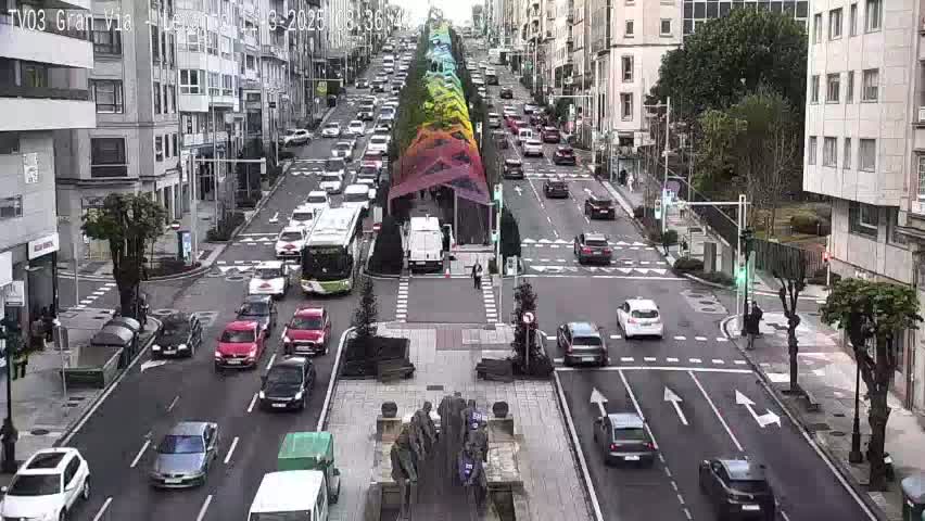 A city street scene on an overcast day shows multiple cars driving in both directions, alongside a colorful, vertical structure in the center of the road.
