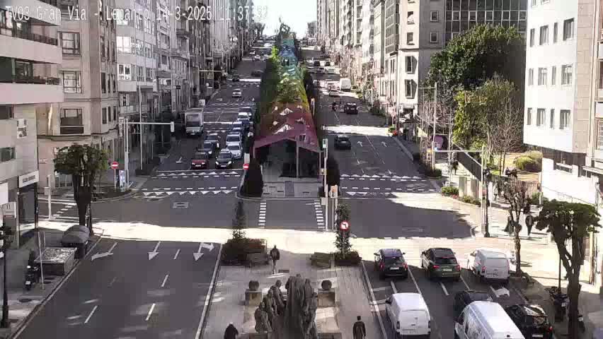 A sunny city street scene shows a multi-lane road intersection with cars, pedestrians, and tall buildings lining the streets, with a central,  shaded area containing a sculpture.