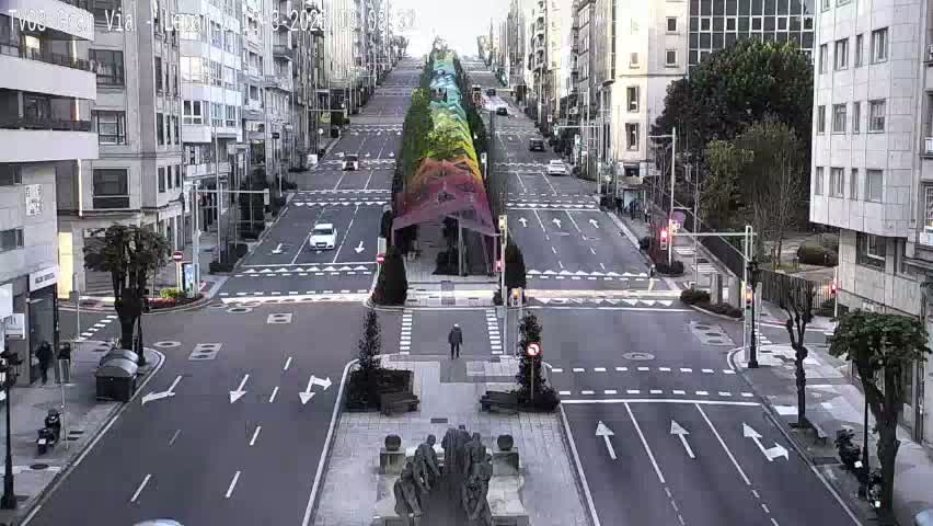 A city street intersection with light traffic under an overcast sky, featuring a colorful structure in the middle of the road and a statue in the center of the intersection.