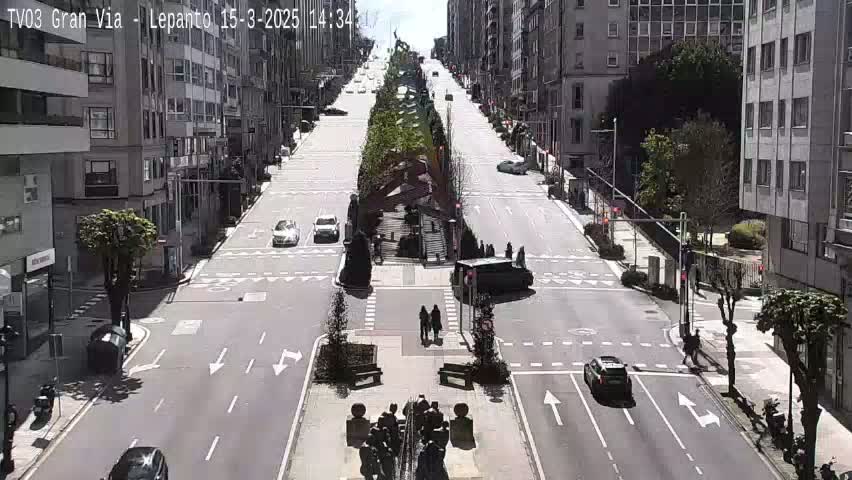 A sunny city street scene shows several cars driving on a wide road that intersects with other streets, with pedestrians crossing and buildings lining the sides.