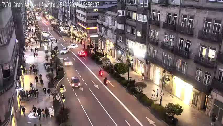 A city street at night, under dark, overcast skies, shows pedestrians walking along the sidewalks and cars driving on a multi-lane road lined with buildings.