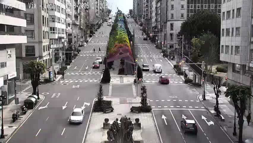 A city street intersection with several cars, a sculpture in the center, and a vibrant, colorful installation lining the median, all under a partly cloudy sky.