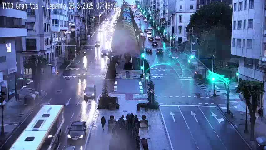 A wet city street scene at night shows traffic moving along a multi-lane road lined with tall buildings and trees, under streetlights.
