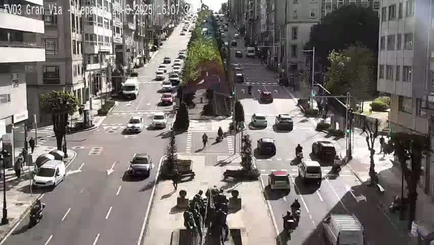 A sunny day on a city street, with numerous cars and pedestrians at an intersection, and buildings lining both sides of the road.