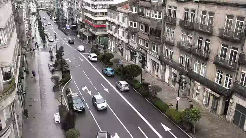 A wet city street with several cars driving along a street lined with multi-story buildings.
