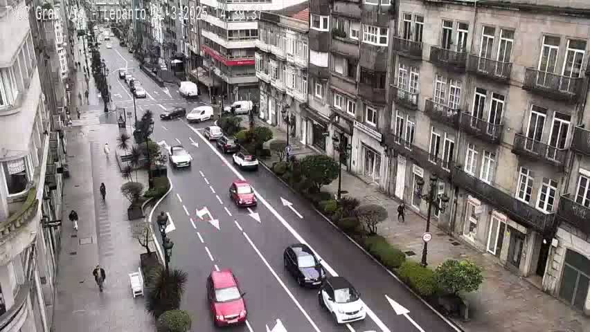 A street scene shows several cars driving on a city street lined with multi-story buildings under an overcast sky.
