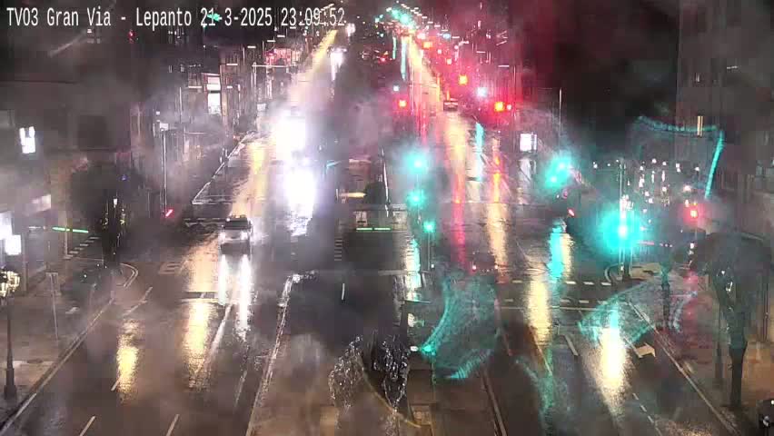 A rain-slicked city street at night shows several vehicles driving through a busy intersection with traffic lights.