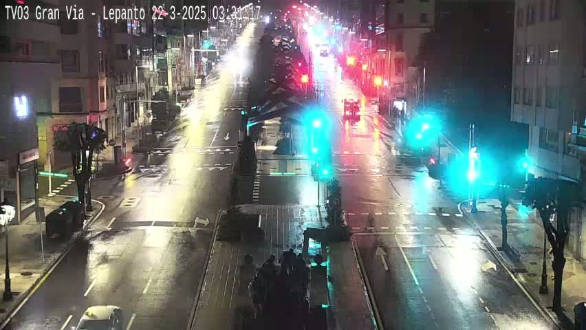 A rain-slicked city street at night is illuminated by streetlights and the headlights of sparsely moving vehicles.