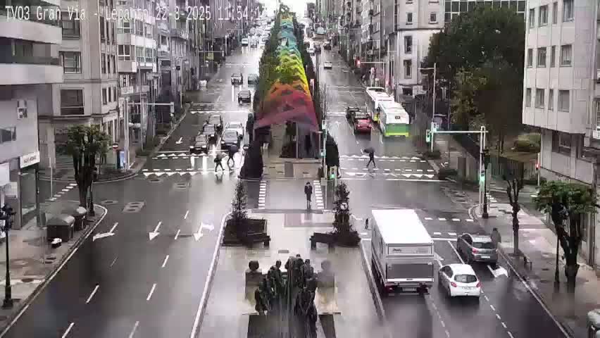A rainy city street scene shows several vehicles driving past multi-story buildings and a colorful, arched structure in the middle of the road.