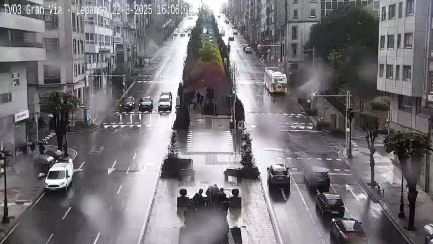 A rainy city street scene shows several cars driving along a wide, wet road lined with buildings and trees.