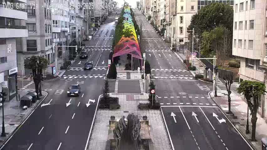 A mostly empty city street with several cars, lined with buildings on either side, features a rainbow-colored canopy structure in the middle, under a cloudy sky.