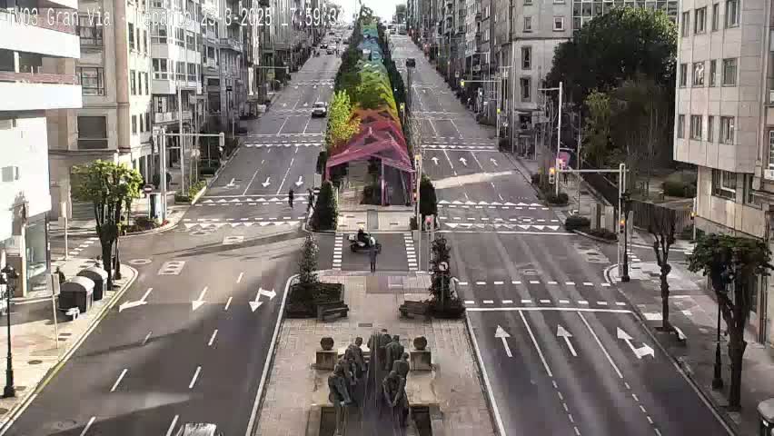 A mostly empty, wide city street with a median featuring colorful plantings and a monument at its center, under a partly sunny sky.