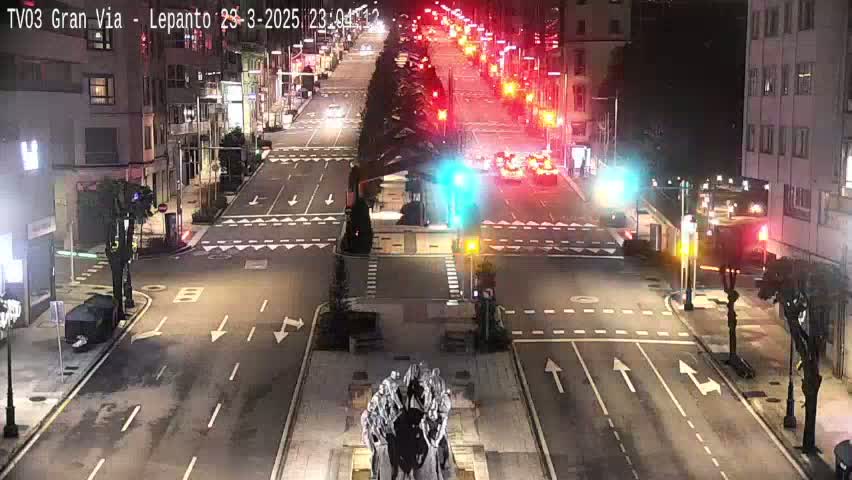A nighttime view of a city intersection with traffic lights and several vehicles driving on a multi-lane road, with a statue in the center of the intersection.