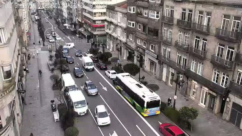 A city street scene on an overcast day shows numerous cars and a bus traveling in both directions, with pedestrians walking along the sidewalks and parked vehicles lining the street.
