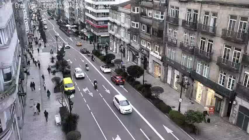 A city street scene on an overcast day shows several cars and pedestrians moving along a multi-lane road that runs between tall buildings.