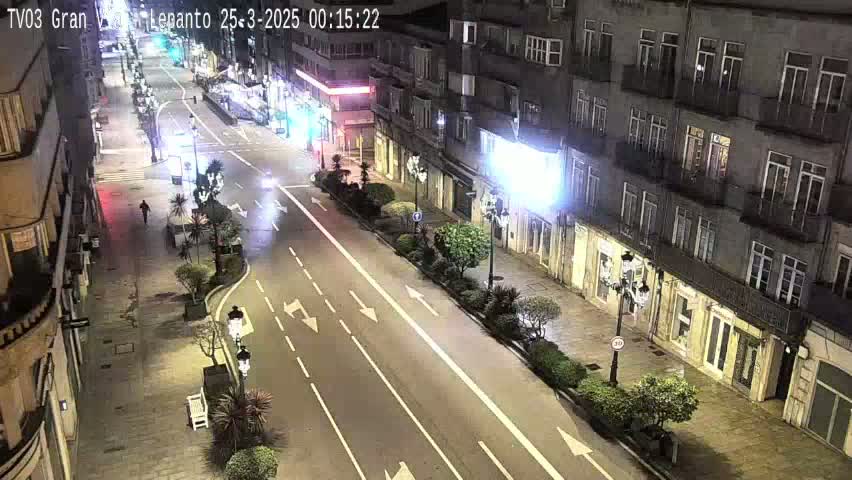 A nighttime street scene shows a mostly empty, multi-lane road lined with buildings and sparsely planted trees.