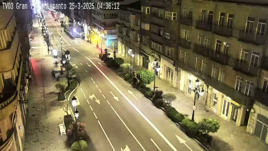 A nighttime, high-angle view of a city street with sparsely spaced vehicles, lined with buildings and streetlights, and small trees and shrubs along the sidewalks.