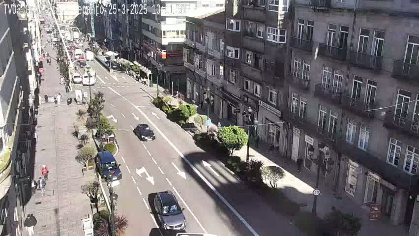 A sunny city street scene shows several cars driving on a multi-lane road, flanked by buildings, pedestrians, and landscaping.