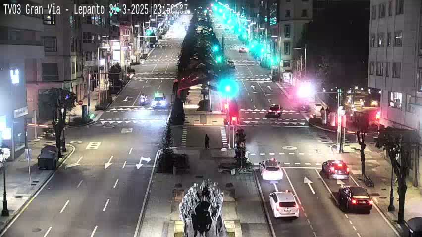 A nighttime view of a city street with traffic flowing in both directions under streetlights, with some vehicles stopped at intersections.