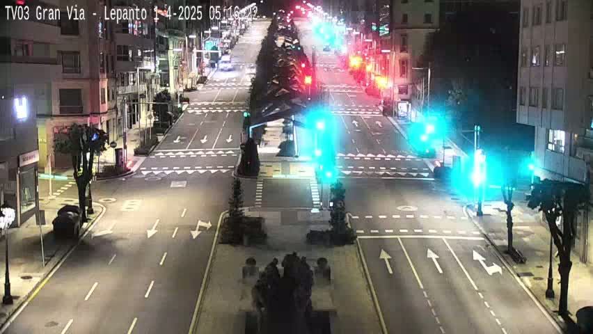 A nighttime, city street scene shows a wide, multi-lane road with traffic lights, sparse traffic, and buildings lining either side.