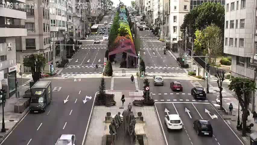 A city street scene with a colorful, abstract art installation in the center, traffic moving in both directions, and pedestrians walking along the sidewalks under a partly sunny sky.