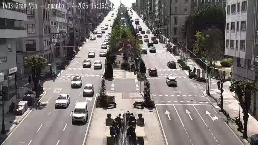 A sunny city street with light traffic, lined with multi-story buildings and a central median with a fountain.