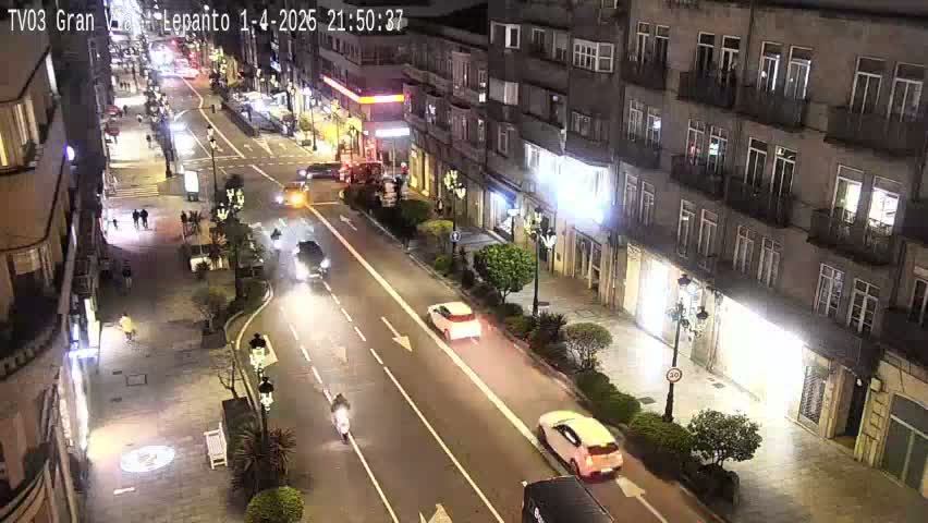 A nighttime street scene shows cars driving on a multi-lane road that passes by a multi-story building with streetlights illuminating the area.