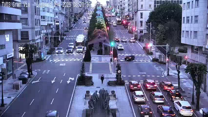 A wide shot of a city street intersection at dusk, with several cars stopped at traffic lights, and a statue in the center of the intersection.