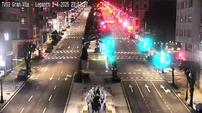 A nighttime, high-angle view of a city intersection with a statue in the center, traffic lights showing green and red, and sparse vehicle traffic on the roads.
