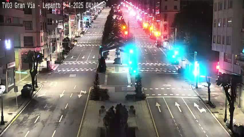 A nighttime, city street intersection with traffic lights showing green and a few cars, surrounded by buildings and trees.