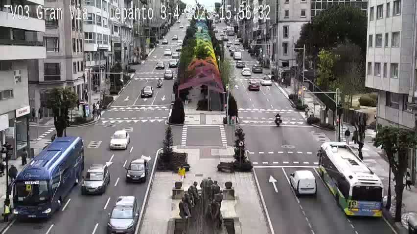 A city street scene on a partly cloudy day shows several cars and buses navigating a multi-lane intersection with a colorful installation in the center.