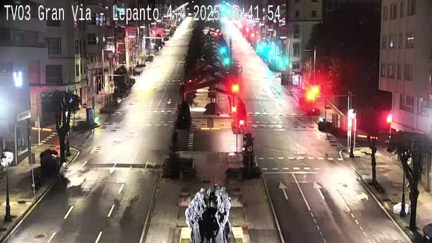 A mostly empty, wet city street at night is illuminated by streetlights and traffic signals, with a large statue visible in the center.