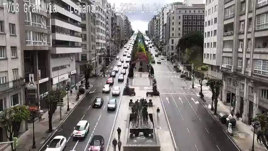 A wet city street scene shows multiple cars driving in both directions on a wide avenue lined with multi-story buildings, and a central median with a modern sculpture.