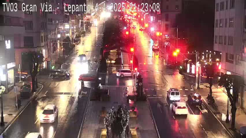 A rain-slicked city street at night is filled with cars moving through intersections under illuminated traffic signals.