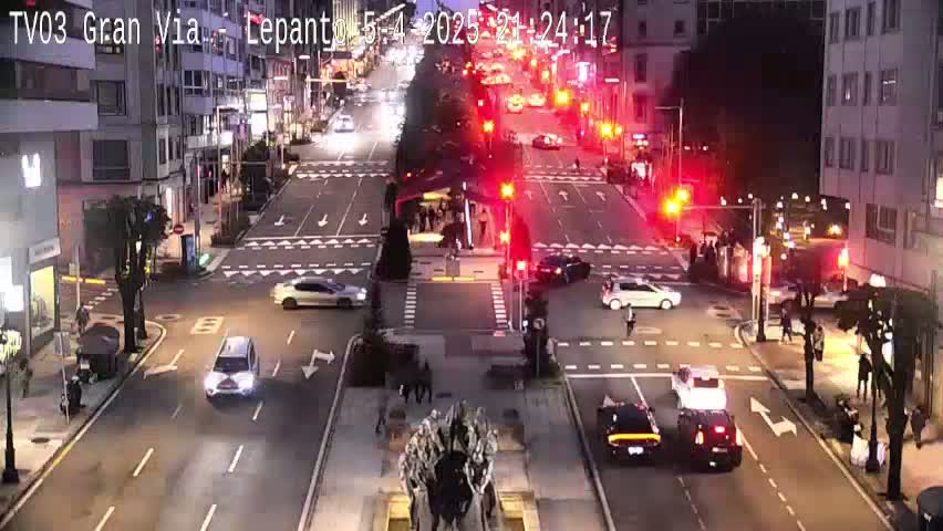 A nighttime city intersection with several cars and pedestrians, under mostly clear skies.