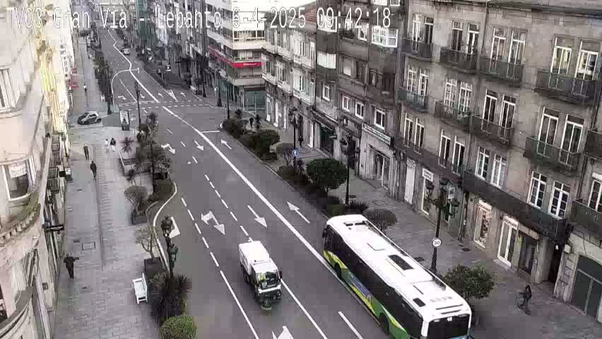 A street scene shows a long, mostly empty road flanked by multi-story buildings under a mostly clear sky, with a street sweeper and a bus driving along the road.