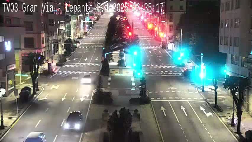 A nighttime, city street intersection with traffic lights, several cars, and buildings lining the streets.