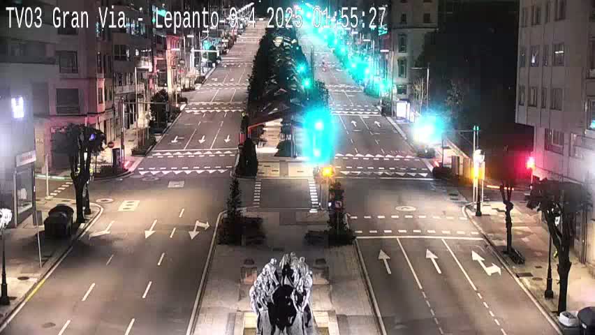 A mostly empty, wide city street at night is illuminated by streetlights, with a large statue in the center and traffic signals at intersections.