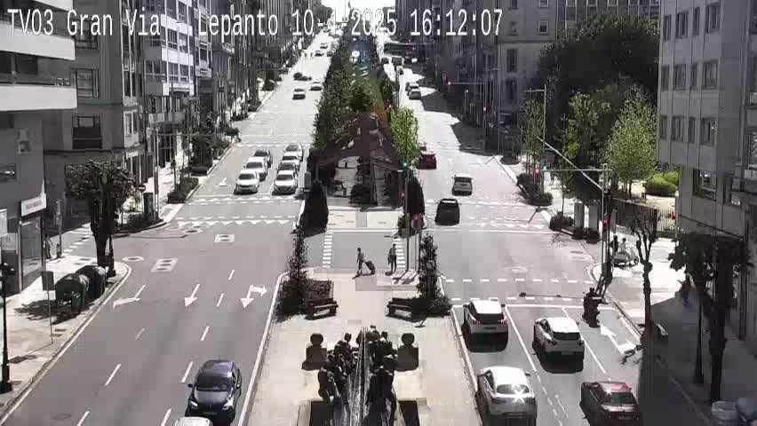 A sunny city street scene shows cars driving on a multi-lane road that intersects with a cross street, and pedestrians walking near a central fountain.