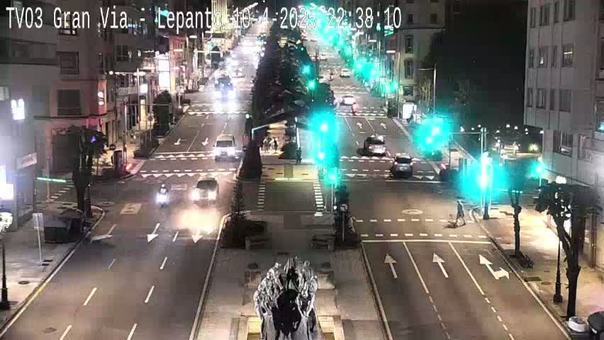 A nighttime, city street scene shows several cars at intersections with traffic signals, and a statue in the center of the road.