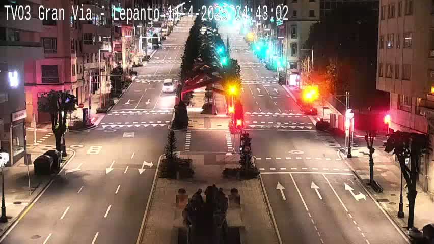 A mostly empty city street at night, illuminated by streetlights and traffic signals, with a few cars visible.