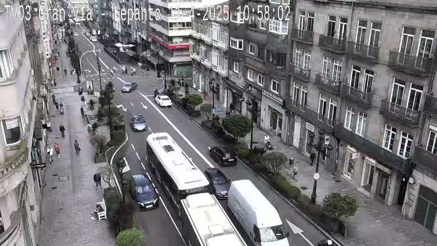 A city street scene on a partly cloudy day shows traffic moving along a wide street lined with multi-story buildings and pedestrians walking on sidewalks.