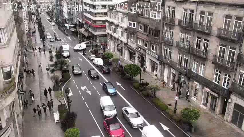 A city street scene shows multiple cars and pedestrians on a multi-lane road lined with buildings under a cloudy sky.