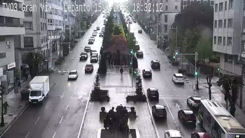 A rainy city street scene shows several cars driving in both directions, with a white van parked on the side of the road and a bus stopped further down.