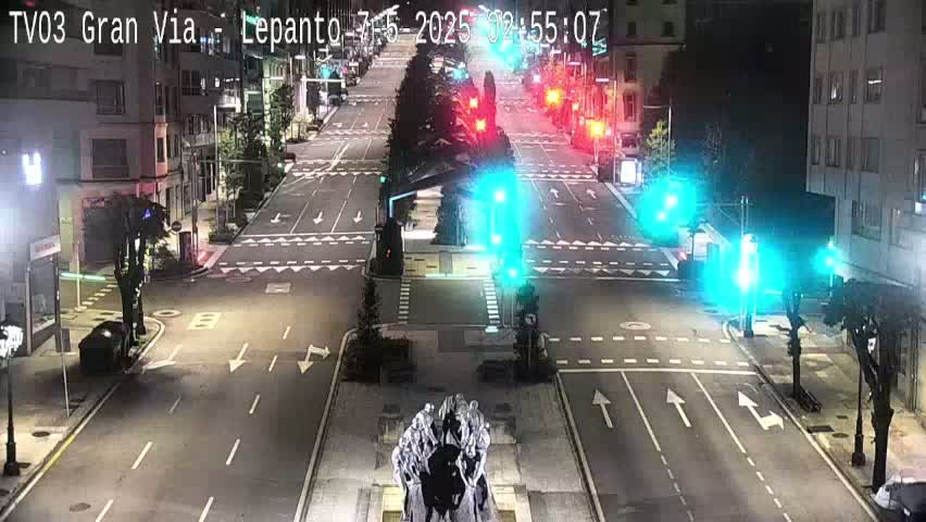 A nighttime, mostly empty street intersection with traffic lights showing green, centered on a statue in a plaza.